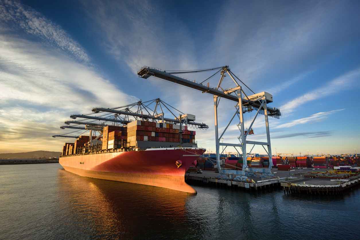 Aerial shot of a massive cargo ship loaded with containers docked in the Port of Long Beach, with the booms of ship-to-shore gantry cranes reaching out from the pier of the terminal. Authorization was obtained from the Port of Long Beach Security Division and POLB Harbor Patrol for this operation in restricted airspace.