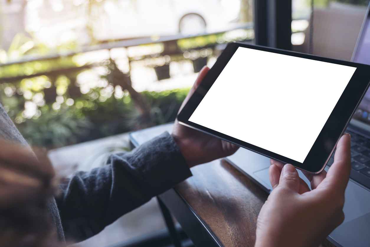 Mockup image of hands holding black tablet pc with white blank screen and laptop on wooden table background in cafe