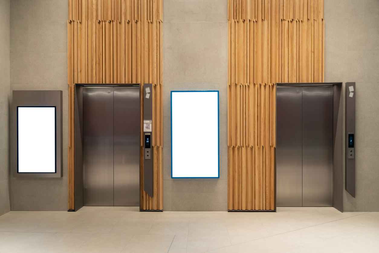 Contemporary elevator lobby featuring two modern elevators with polished metal doors on corridor lobby, mockup billboard with frame between elevators, blank space for advertisement or announcement