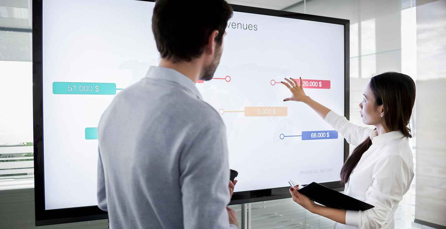 Male and female colleague standing by large screen in meeting room, going over financial presentation shown on screen in meeting room.
