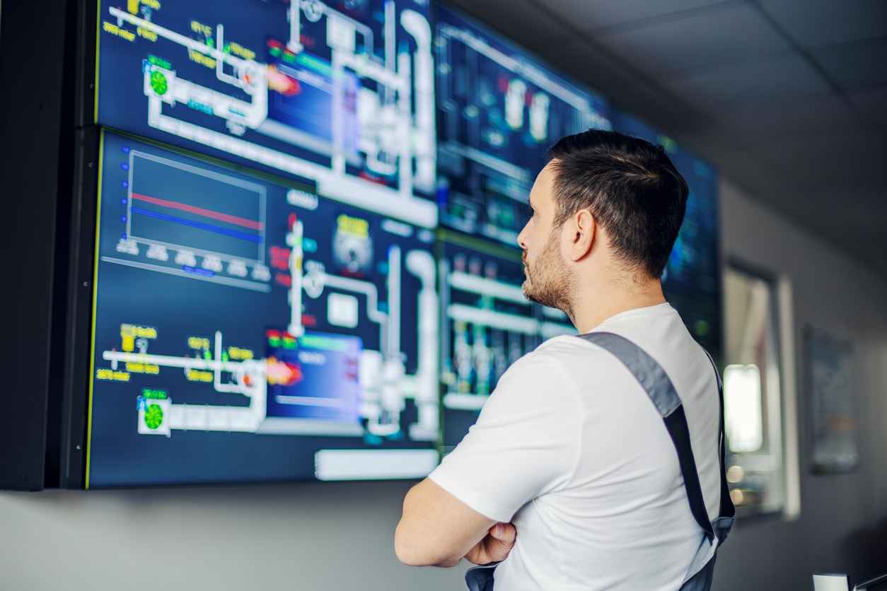 Male engineer stands with arms crossed in a modern control room, monitoring large screens showing data visualizations, graphs and schematics of industrial automation systems